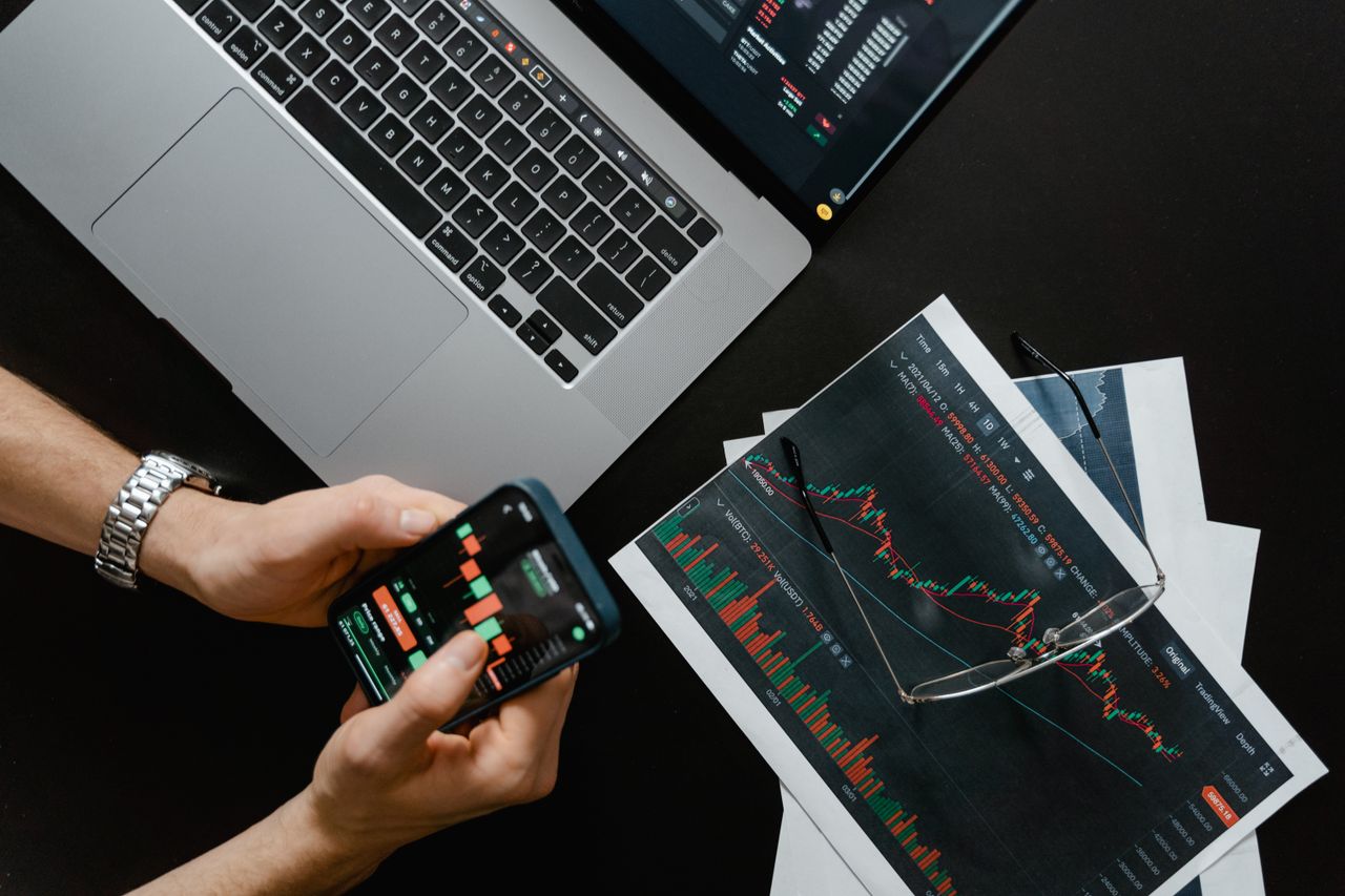 about-us-01 Hands using smartphone beside laptop with stock charts, showcasing digital trading.